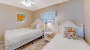 A bedroom with two single beds, a ceiling fan, a nightstand with a lamp, and a window with blinds. - Luxury, oceanfront condo with outdoor pool & patio - snowbird-friendly (Miramar Beach)