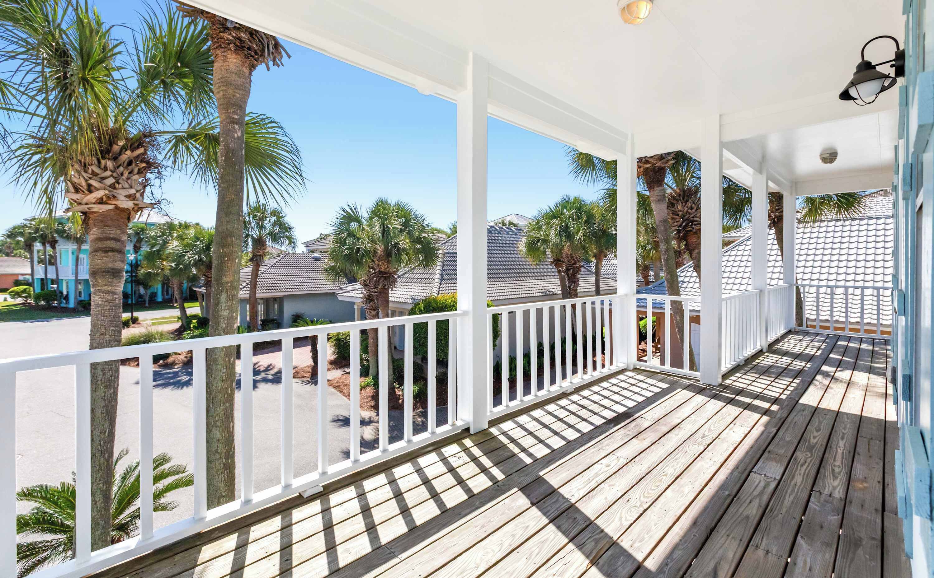 A covered wooden deck with white railings, palm trees, and houses in the background.
