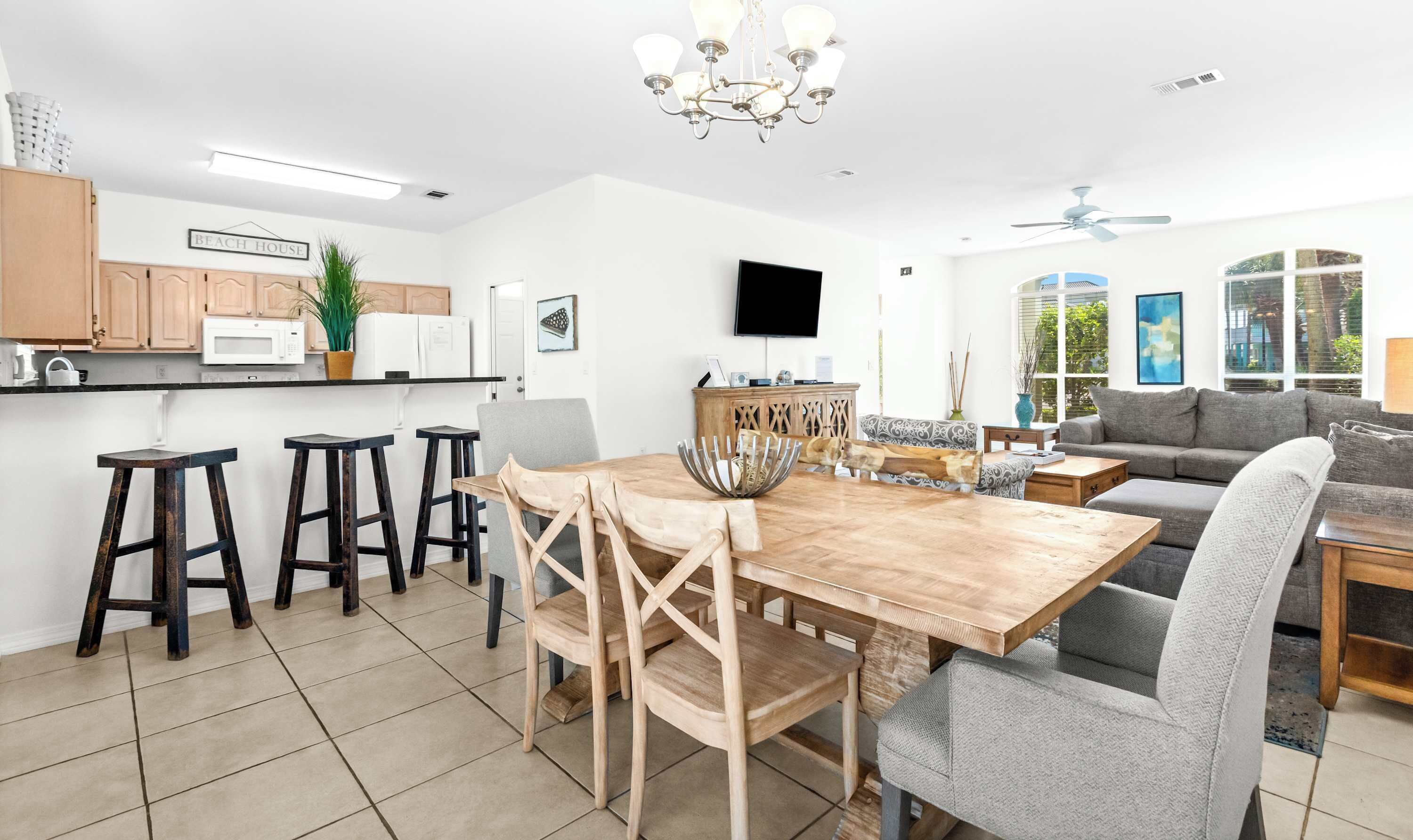 A modern kitchen with wooden cabinets, a dining area with a wooden table and chairs, a grey sofa, and a TV mounted on the wall.