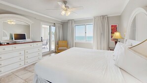 Une chambre spacieuse avec un grand lit, un ventilateur de plafond, un téléviseur posé sur une commode et une vue sur l’océan par les fenêtres.