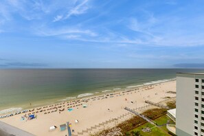 A beach with umbrellas and people, a building with a balcony, and a clear sky. - 11th-floor, Gulf-front condo with views, pools, hot tub & tennis (Perdido Key)