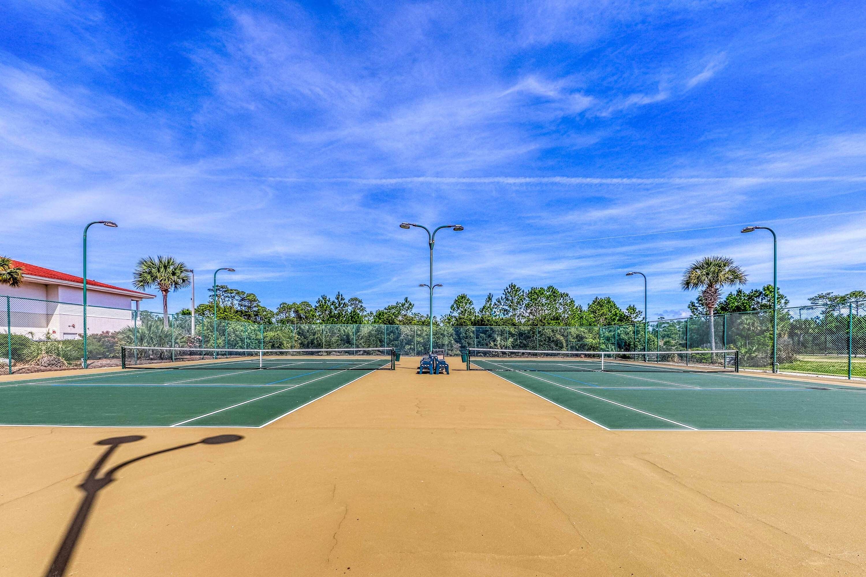 A tennis court with a net, green and brown surface, and street lamps.