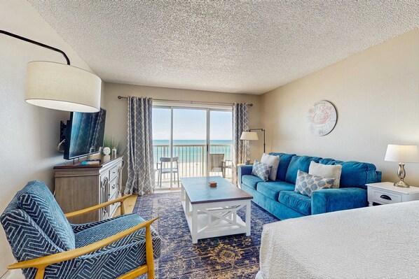 A room with a blue sofa, a wooden chair, a white coffee table, and a TV on a cabinet. There is a balcony with curtains and a view of the sea. - Fourth floor oceanfront studio, balcony, full kitchen, heated outdoor shared pool, snowbirds welcome (Fort Walton Beach)