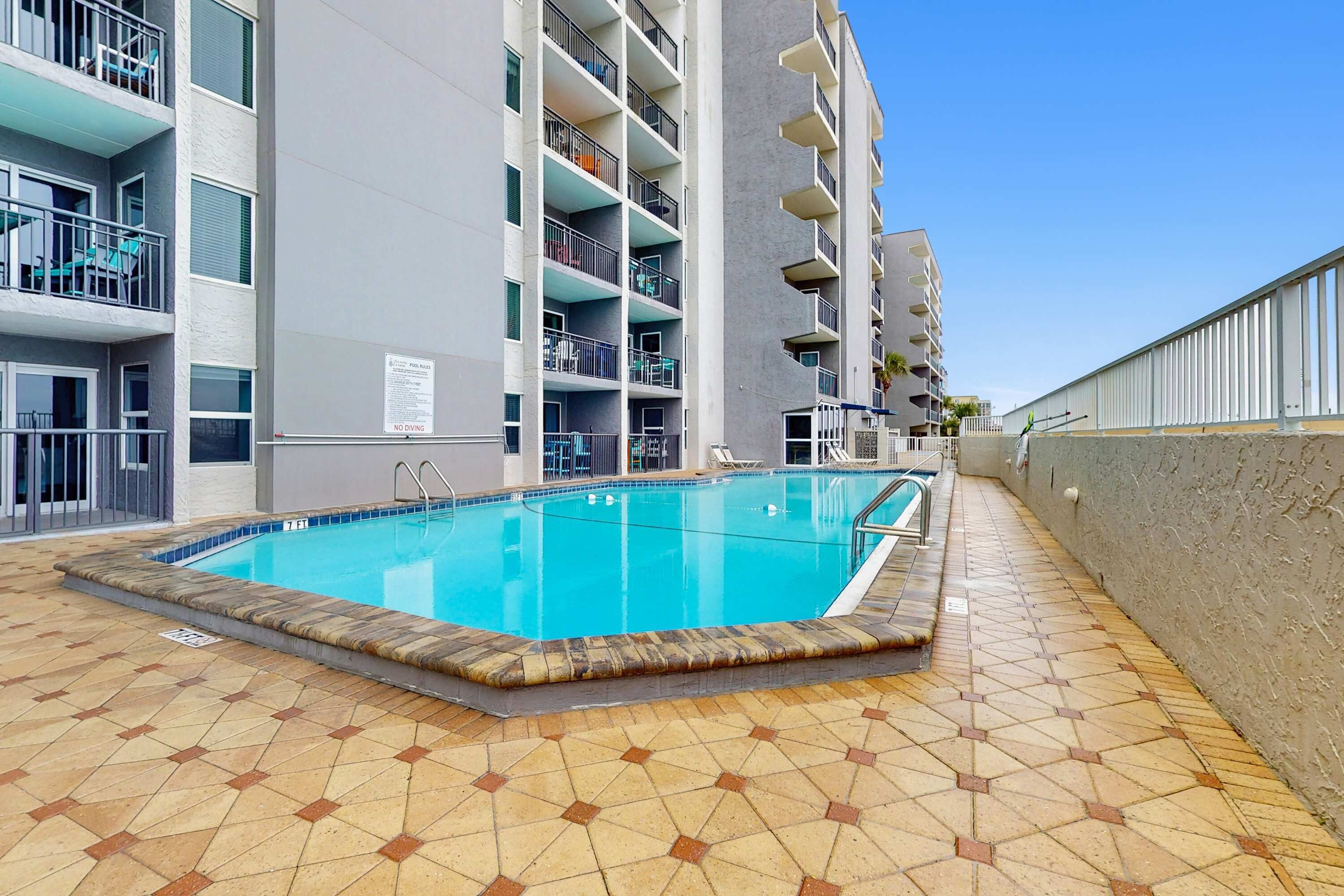 Piscina adjacente a um edifício com várias andares, varandas e chão em azulejo.