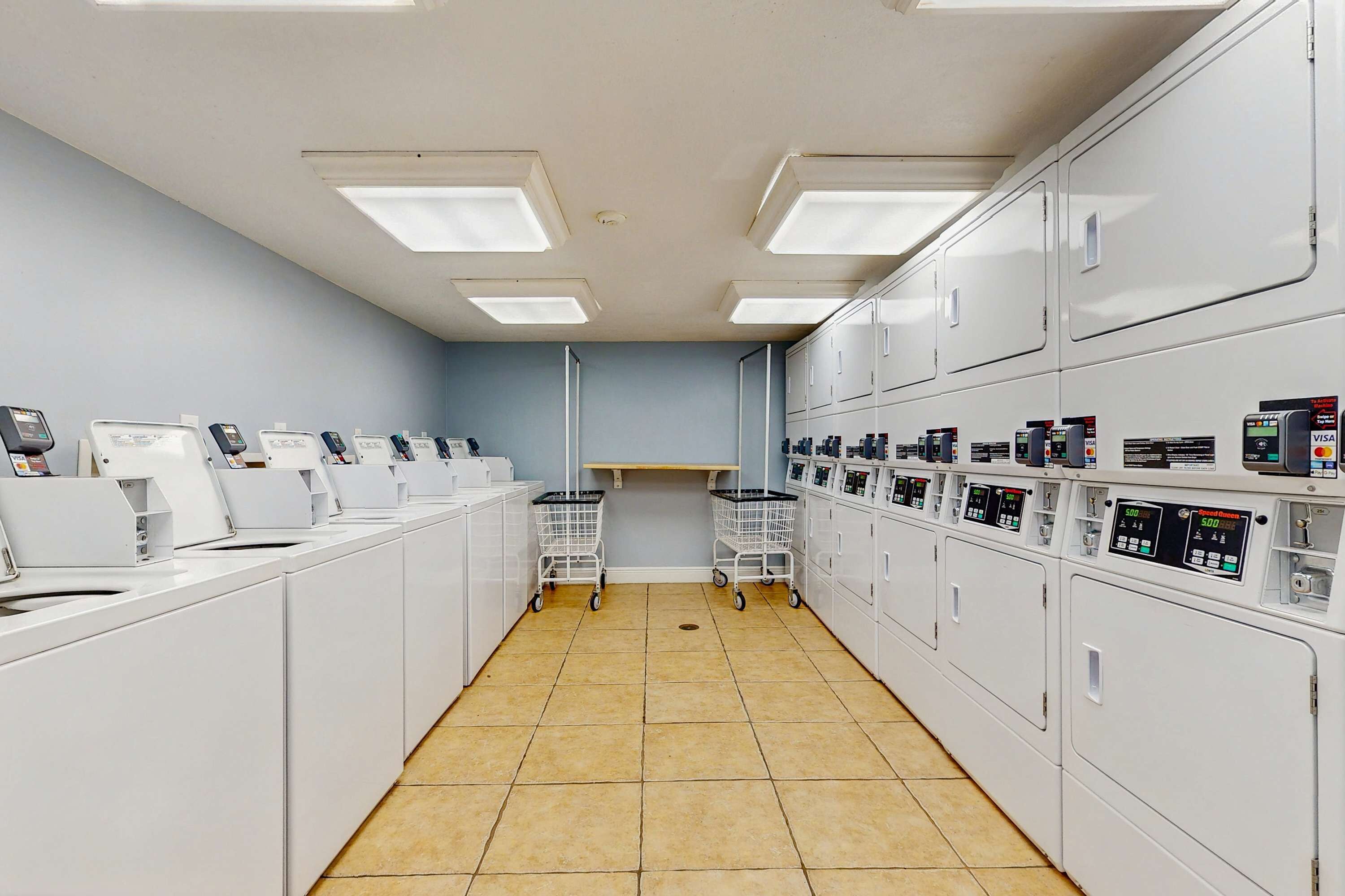 Une laverie équipée de plusieurs machines à laver et de plusieurs sèche-linge, avec une allée centrale et un mur de couleur bleu clair.