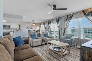 A living room with a sofa, armchairs, a coffee table, and a ceiling fan. There is a kitchen area with cabinets and a microwave. - Beachfront condo with gulf views, pool, gym, & private balcony (Fort Walton Beach)