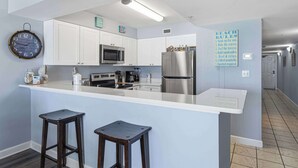 A modern kitchen with white cabinets, a stainless steel refrigerator, a microwave, and a clock on the wall. - Gulf front condo w/ beach setup included - near entertainment (Fort Walton Beach)