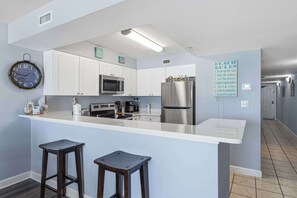 A modern kitchen with white cabinets, a stainless steel refrigerator, a microwave, and a clock on the wall. - Gulf front condo w/ beach setup included - near entertainment (Fort Walton Beach)