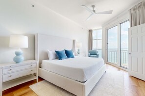A modern bedroom with a large bed, white nightstands, a ceiling fan, and a view of the ocean through large windows. - 18th-floor condo with amazing views, beach access, pools & gym (Perdido Key)