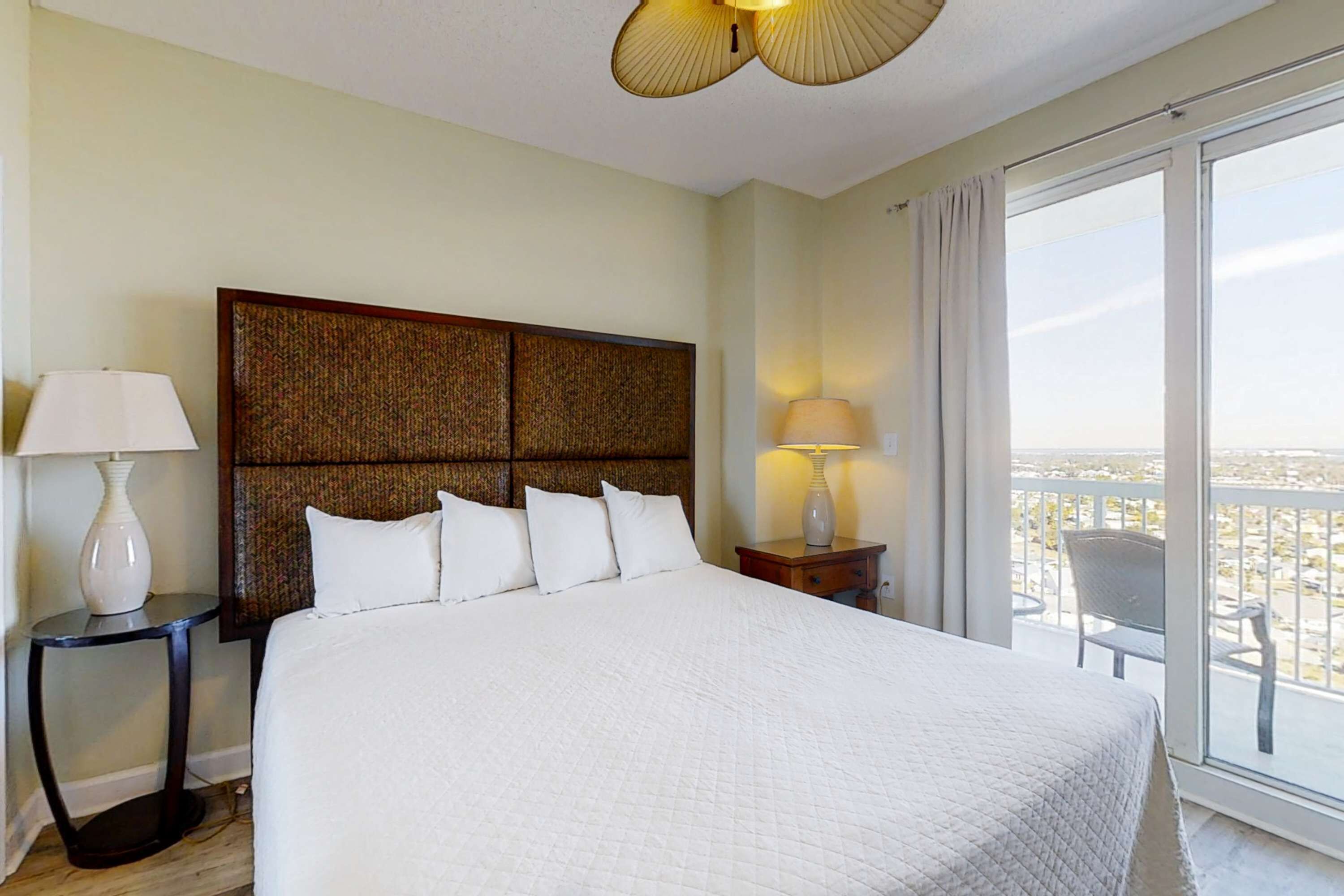 Habitación de hotel con una cama grande, dos mesitas de noche con lámparas y vistas a la ciudad desde un balcón.
