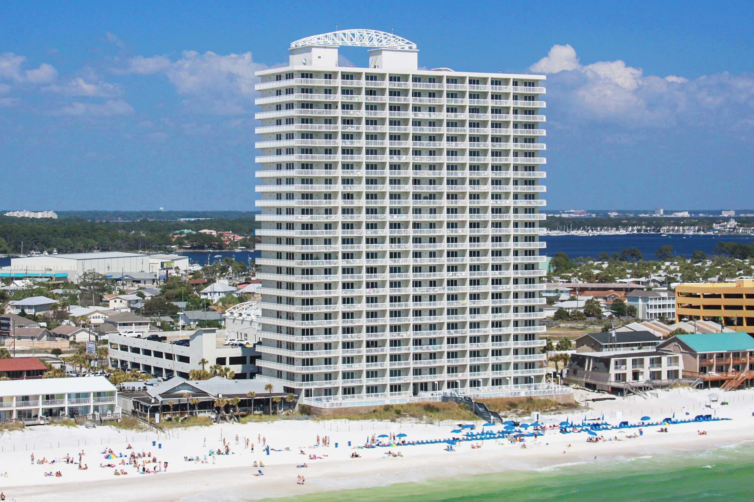 A tall hotel with multiple floors, a beach in the foreground, and a view of the ocean and other buildings in the background.