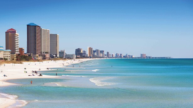 Ein Strand mit klarem türkisblauem Wasser und einer Reihe hoher Gebäude im Hintergrund.