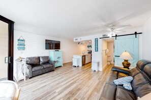 A modern living room with a sofa, a coffee table, and a dining area with a kitchen in the background. - Beachfront condo with a private balcony, three pools, tennis, & picnic area (Panama City Beach)