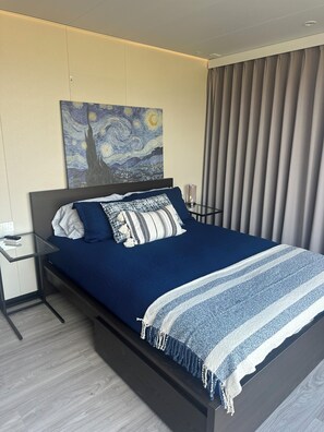 Un dormitorio moderno con una cama, una mesita de noche, un cuadro y un ventanal con cortinas.