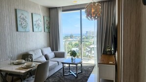 A modern living room with a sofa, dining table, and coffee table. There are framed floral artworks on the wall, a large window with a view, and a decorative hanging light fixture. - 1 BR 5mins to beach Sea view Riviera Wongamat (Muang Pattaya)