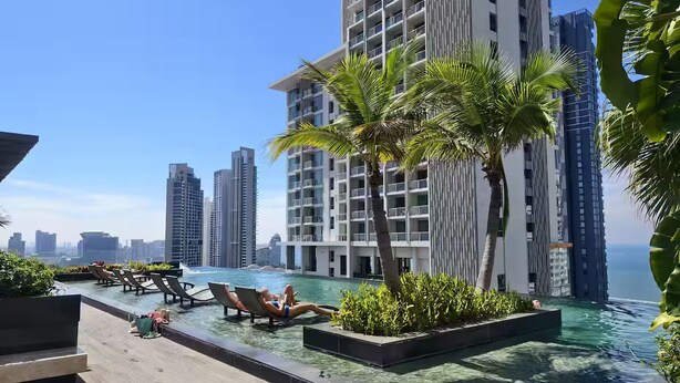 A rooftop infinity pool with lounge chairs, palm trees, and high-rise buildings in the background. - 1 BR 5mins to beach Sea view Riviera Wongamat (Muang Pattaya)