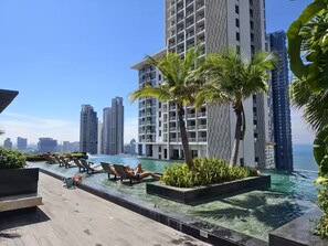A rooftop infinity pool with lounge chairs, palm trees, and high-rise buildings in the background. - 1 BR 5mins to beach Sea view Riviera Wongamat (Muang Pattaya)