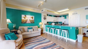 A living room with a sofa, a coffee table, and a teal accent wall. There is a kitchen area with a bar and stools. - 22nd-floor beachfront condo with Gulf views, hot tub, pools, sauna, gym, balcony (Panama City Beach)