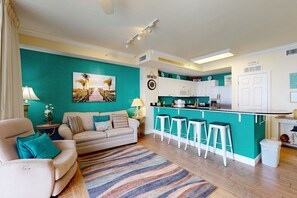 Ein Wohnzimmer mit einer Couch, einem Couchtisch und einer Wand in Türkis. Es gibt eine Küchenzeile mit einer Bar und Hockern.
