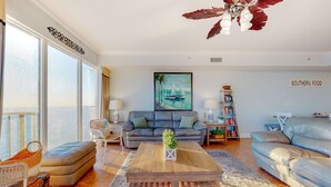 A spacious living room with a large window, a sofa, a coffee table, and a bookshelf. - Spacious high-rise condo with stunning beachfront views, pools & hot tubs (Panama City Beach)
