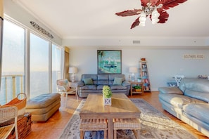 A spacious living room with a large window, a sofa, a coffee table, and a bookshelf. - Spacious high-rise condo with stunning beachfront views, pools & hot tubs (Panama City Beach)
