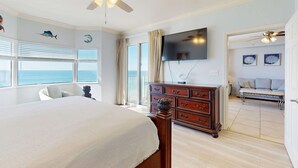 Un dormitorio con una cama grande, un ventilador de techo, un aparador, un televisor y vistas al océano a través de las ventanas.
