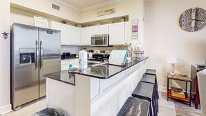 A modern kitchen with a stainless steel refrigerator, a microwave, and a coffee maker. There is a clock on the wall and a small table with a lamp. - Exquisite Gulf front condo w/shared pool, hot tub &, gym/Roman spa on-site (Panama City Beach)