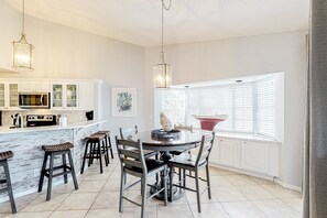 A modern kitchen with white cabinetry, a central island with stools, a dining table with chairs, pendant lights, and a view through a window with blinds. - Dog-friendly condo with a shared, outdoor pool & fast WiFi - steps from beach (Miramar Beach)