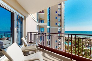 Un balcón con sillas y una mesa blancos, con vista a una playa y una piscina.
