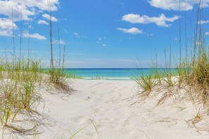Une plage de sable blanc, avec de hautes herbes au premier plan, qui s’étend jusqu’à une mer bleue et limpide sous un ciel partiellement nuageux.