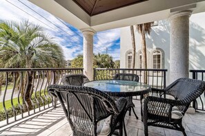 A covered outdoor patio with a round glass table and wicker chairs, surrounded by palm trees and a building with arched windows. - Spacious Beach Home with Pool & Balconies - Short Walk to Private Beach Access (Miramar Beach)