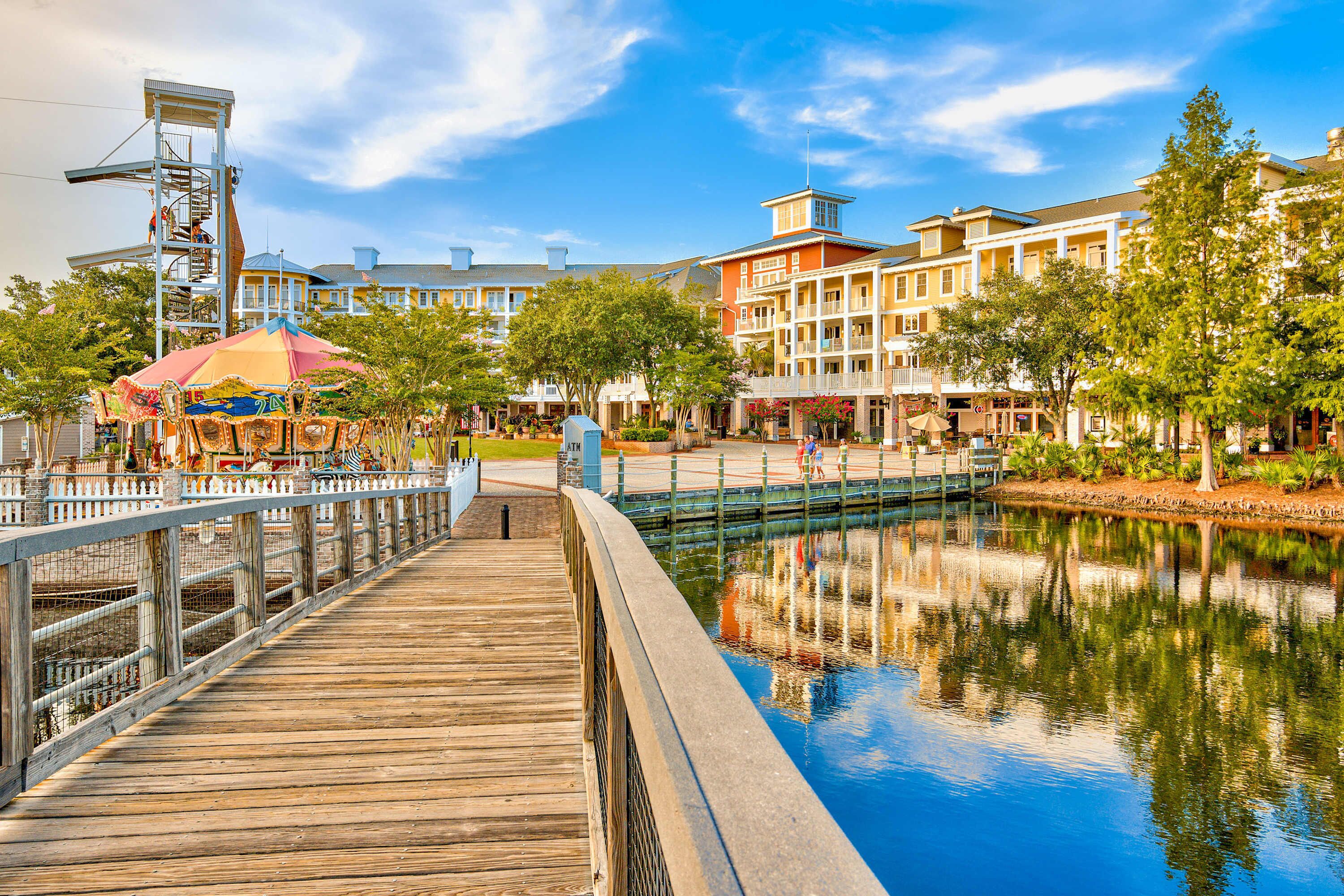 A wooden boardwalk over a calm water body with a carousel and buildings in the background.