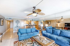 A spacious living room with blue sofas, a coffee table with a decorative item, a ceiling fan, and a large mirror reflecting the kitchen area. - Seventh-floor oceanfront condo with pool access, ocean view, & central AC (Fort Walton Beach)