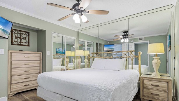 Un dormitorio con cama, mesitas de noche, cómoda, ventilador de techo y televisor.