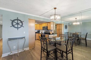 A modern kitchen with wooden cabinets, a central island, and a dining area with a glass table and metal chairs. - Seventh-floor oceanfront condo with pool access, ocean view, & central AC (Fort Walton Beach)