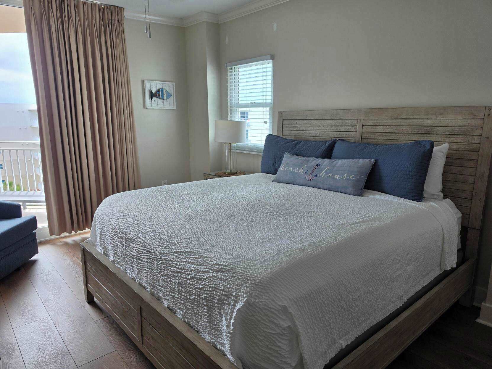 A bedroom with a large bed, a wooden headboard, a nightstand, a lamp, a window with blinds, and a view of the outdoors.
