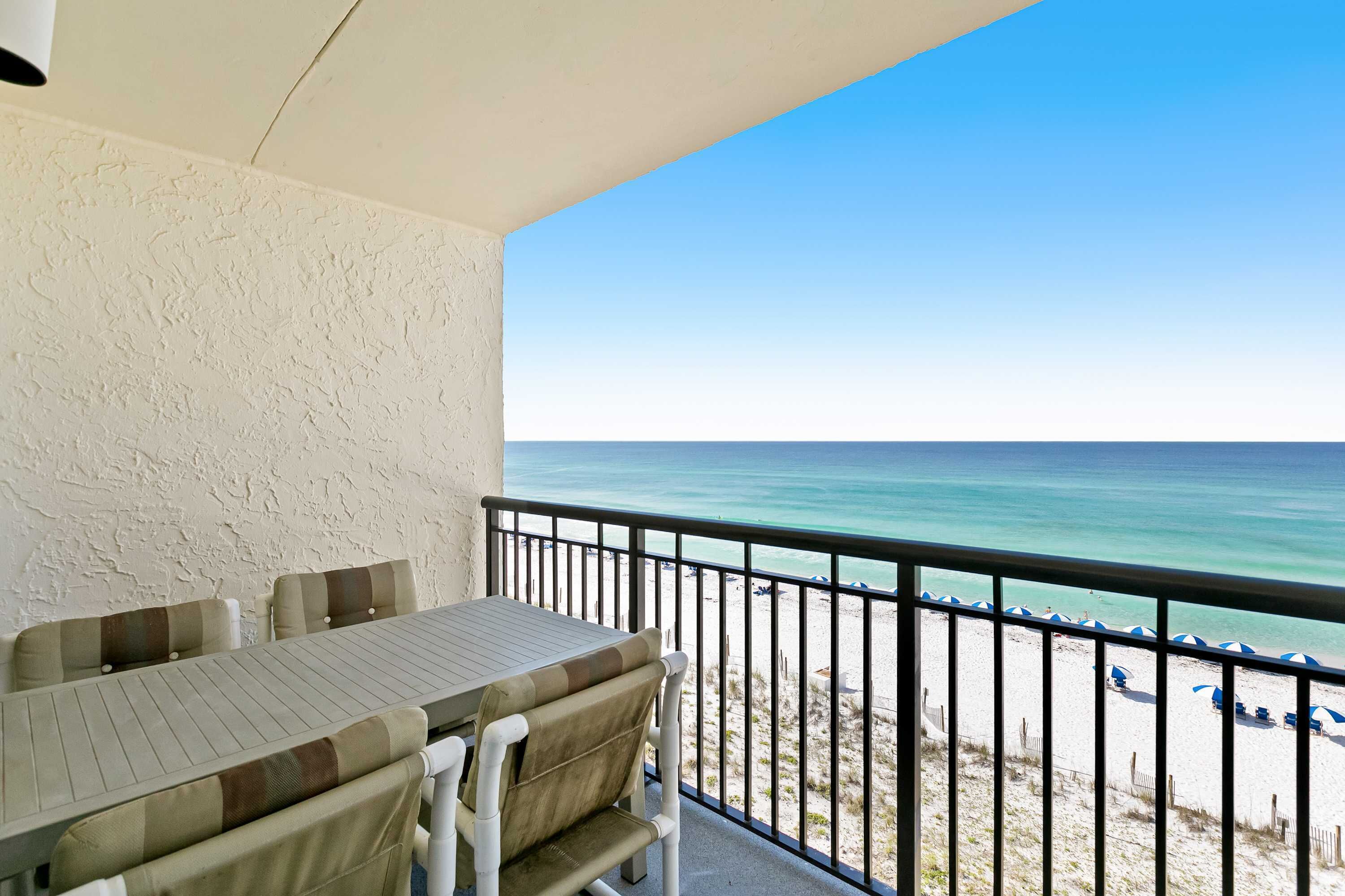 Un balcon avec une table et des chaises, donnant sur une plage parsemée de parasols et bordée d’une clôture.