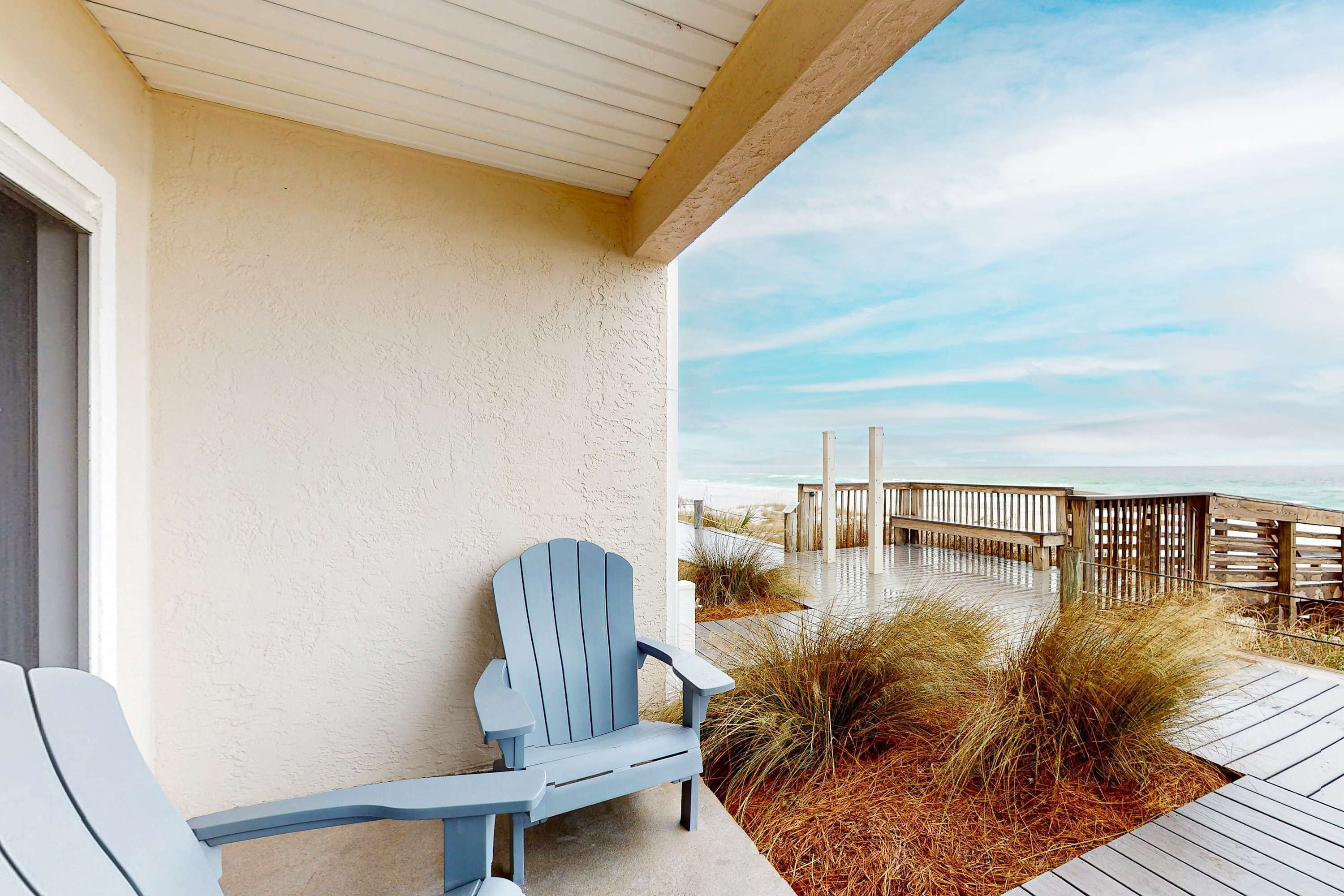 Eine Holzterrasse mit einem blauen Adirondack-Sessel, einem Holzzaun und Blick auf den Strand.