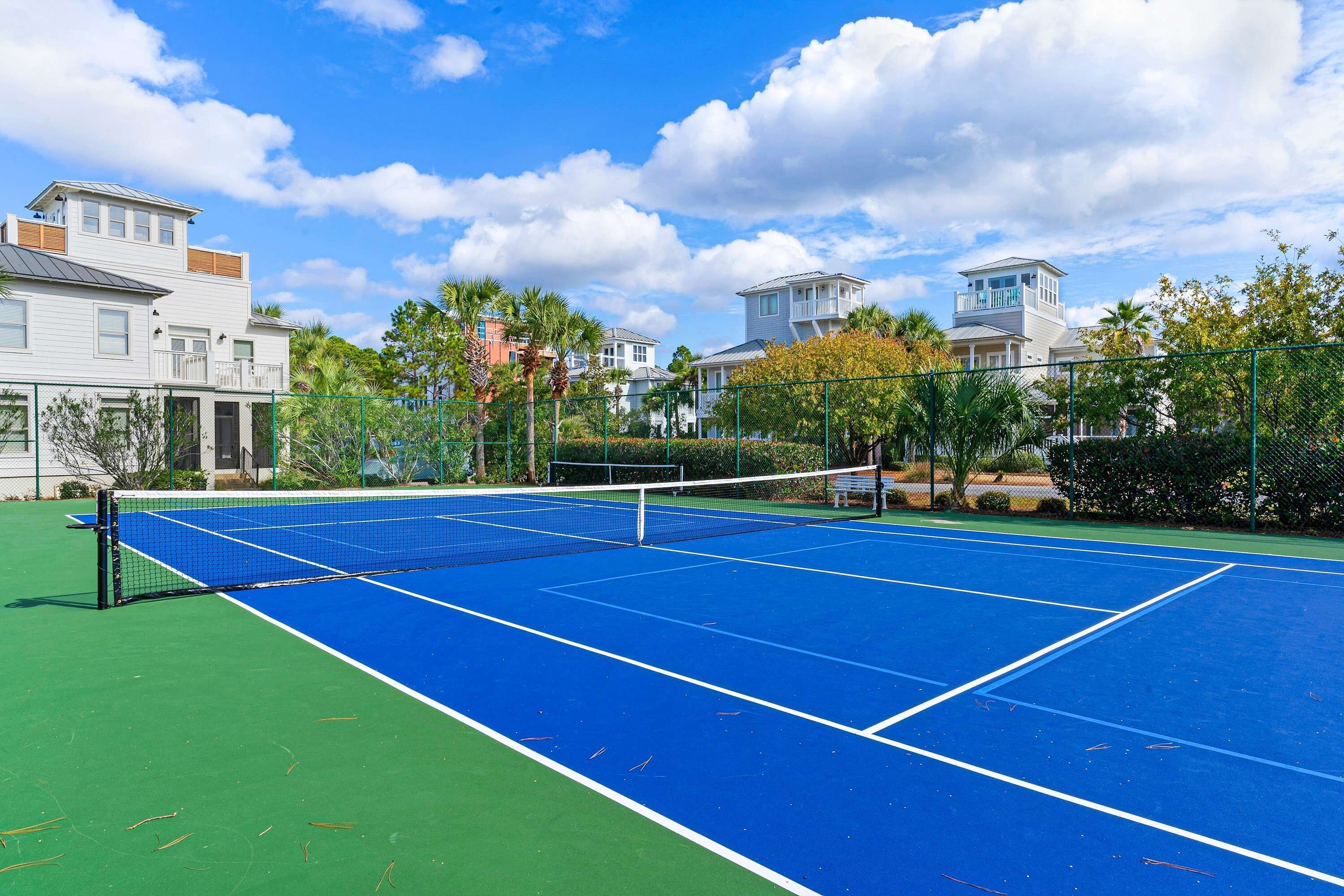 A tennis court with a net, surrounded by green fencing and palm trees.