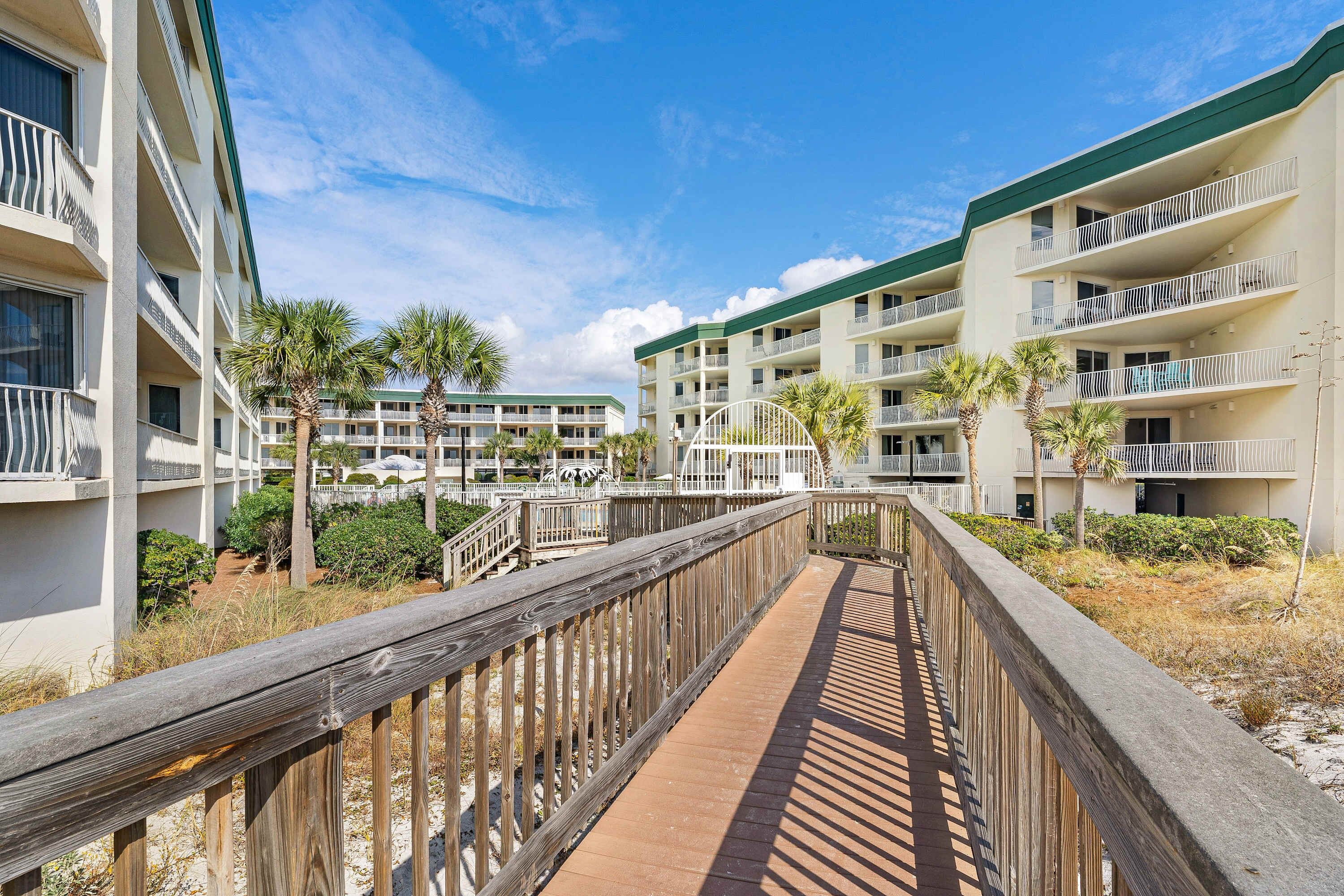 A wooden boardwalk leading to a multi-story hotel with balconies and palm trees.