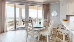 Un comedor con una mesa grande y sillas, vistas a la playa y una zona de cocina con mostrador y armarios.