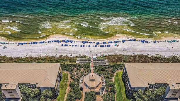 Vista aérea de un resort en la playa con un edificio grande en el centro, rodeado de palmeras y una playa de arena con sombrillas.