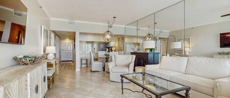 A spacious living room with a white sofa, a glass coffee table, and a dining area in the background.