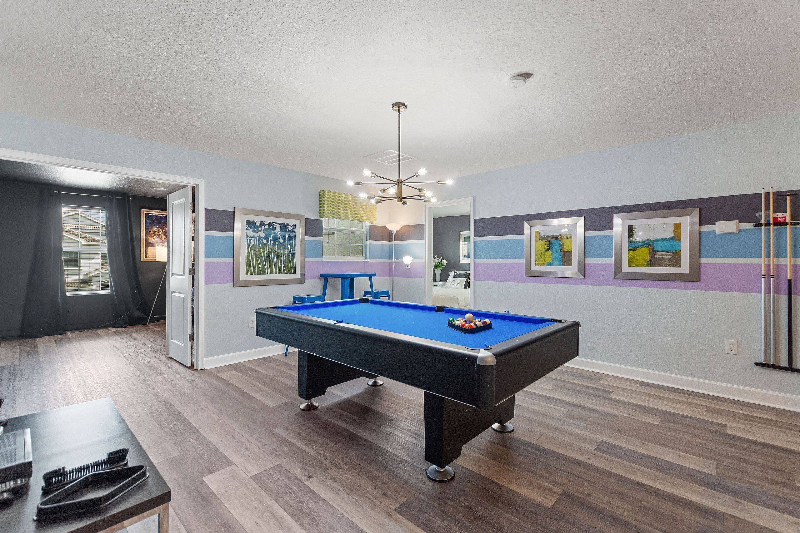 A pool table in a room with wooden flooring, a ceiling light fixture, and framed artwork on the walls.