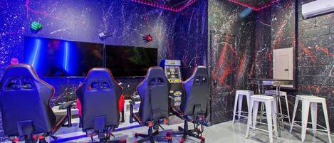 Una sala de jocs amb quatre seients gamer, un monitor gran i un panell de control.