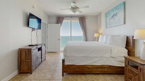 A bedroom with a bed, a TV, a ceiling fan, a painting, and a view of the outside. - 16th-floor oceanfront condo with ocean view, WiFi, and hot tub/pool (Panama City Beach)