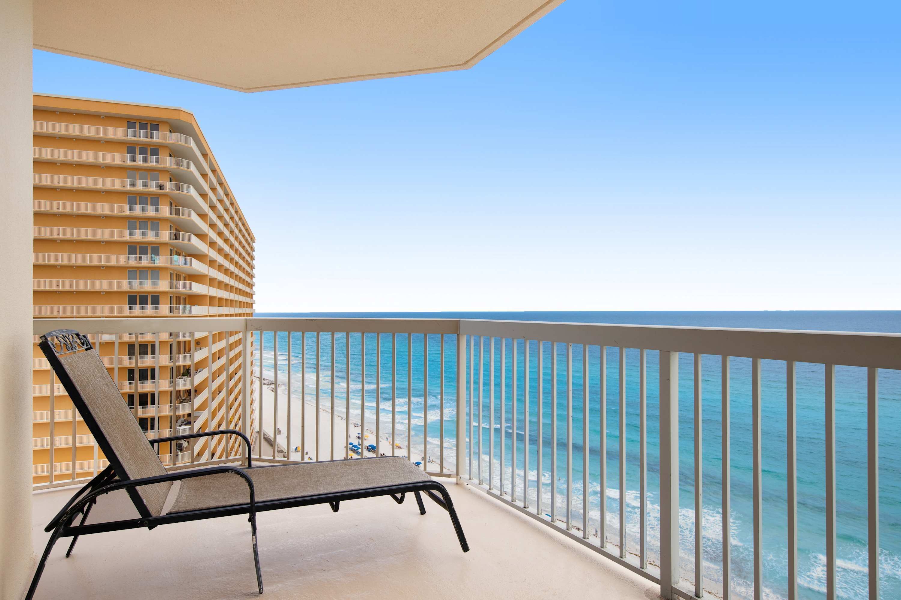 A balcony with a lounge chair, overlooking a beach and a hotel building.
