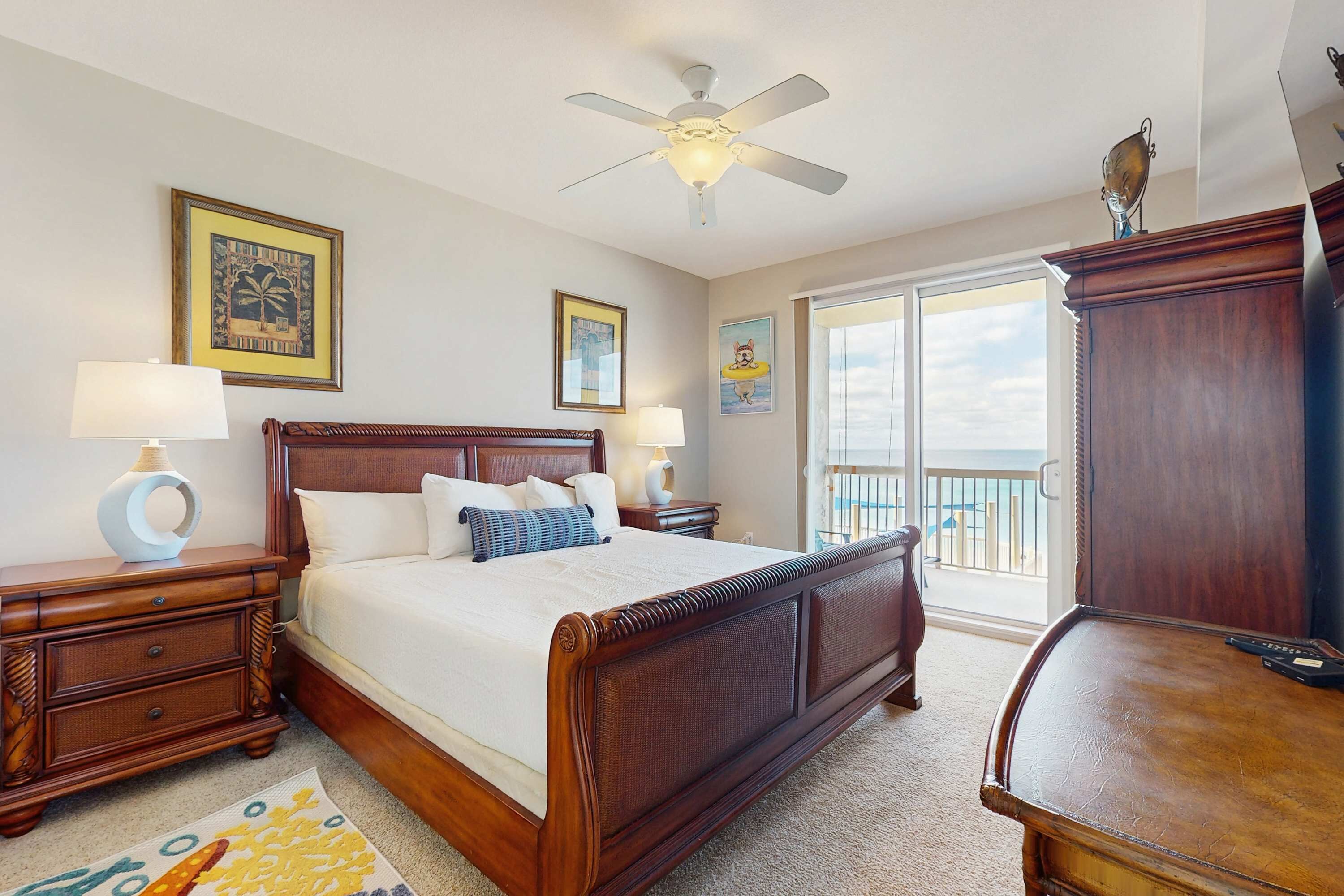 A bedroom with a large bed, wooden headboard, two bedside tables with lamps, a ceiling fan, and a view of the outdoors through a sliding door.