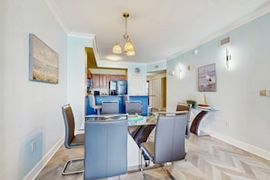 A modern kitchen with blue cabinets and a central island, a dining area with a glass table and grey chairs, and a hallway leading to a door. - Sand-front condo with pool, hot tub, gym, scenic balcony, smart TVs, W/D, & AC (Panama City Beach)