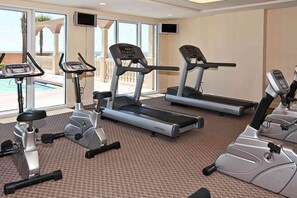 A gym with treadmills and stationary bikes, large windows with a view of a pool and palm trees. - Sand-front condo with pool, hot tub, gym, scenic balcony, smart TVs, W/D, & AC (Panama City Beach)